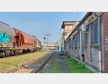 Bahnhofsgelände mit Gleisen und abgestellten Güterwaggons auf der linken Seite, daneben ein längliches, graues Gebäude mit vergitterten Fenstern und Klimaanlagen an der Fassade; im Hintergrund weitere Gleise und Masten.