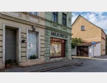 Historische Straßenszene mit einem zweistöckigen Gebäude in grünlicher Farbgebung, das die Aufschrift "Wilh. Riese" trägt und eine Schusterwerkstatt aus dem Jahr 1790 beherbergt. Rechts daneben ein gelbes Wohnhaus, ein gepflasterter Gehweg und eine kleine Seitengasse.
