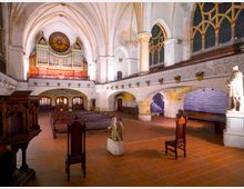 Innenraum einer historischen Kirche mit gotischen Gewölbedecken, großen verzierten Fenstern und einer auffälligen Kirchenorgel auf der Empore. Im Vordergrund stehen Holzstühle und Statuen auf einem Boden aus roten Fliesen, während Bänke und Bögen den Raum strukturieren.