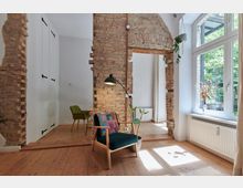 Wohnraum mit freigelegten Backsteinwänden, großen weißen Fenstern, Holzboden und einem gemütlichen Stuhl mit Stehlampe im Vordergrund. Im Hintergrund befindet sich ein Bereich mit Esstisch und grünem Stuhl.