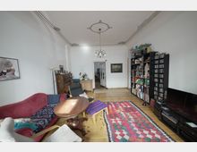 Wohnzimmer mit Holzboden und klassischer Einrichtung. Zu sehen sind ein rotes Sofa, ein runder Holztisch, ein Bücherregal, ein Klavier und ein Teppich mit buntem Muster; die Decke ist mit verzierten Stuckelementen gestaltet.