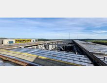 Weitläufige Industrie-Dachkonstruktion mit überwiegend metallischen Elementen und sichtbaren Trägern, im Hintergrund ein beige-graues Gebäude mit gelben Verstrebungen; dahinter hügelige Landschaft unter klarem Himmel.
