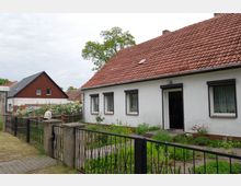 Ein eingeschossiges Wohnhaus mit weißer Fassade und rotem Ziegeldach, umgeben von einem Garten mit Blumen und Sträuchern. Im Vordergrund ist ein Metallzaun mit Tor sowie ein gepflasteter Weg zu sehen; im Hintergrund stehen weitere Häuser.