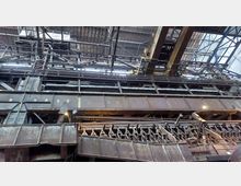 Industriehalle mit massiven Stahlträgern, Rohrleitungen und Oberlicht. Der Raum zeigt eine komplexe Metallkonstruktion mit rostigen Elementen und mechanischen Anlagen, umgeben von einer hohen Decke mit sichtbaren Stahlverstrebungen.