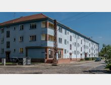 Mehrstöckiges Wohngebäude in hellblauer Farbe mit roten Dachelementen und einigen Backstein-Details an den Ecken und Eingangsbereichen. Der Gehweg und eine Kopfsteinpflasterstraße verlaufen entlang der Front, im Vordergrund stehen Müllcontainer und eine Metalllaterne.