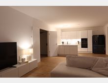 Modernes Wohnzimmer mit offener Küche. Weiße Einbauküche mit schwarzen Geräten, Holzfußboden, ein Sofa im Vordergrund und ein Fernseher auf einem weißen Lowboard links im Bild.