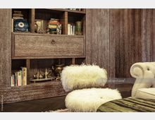 Holzvertäfelter Wohnraum mit einem rustikalen Regal, das Bücher, Deko-Objekte und Teekannen aus Metall enthält. Im Vordergrund ein flauschiger Stuhl mit weißem Kunstfell und ein beigefarbener Sessel mit Nieten, daneben ein runder Tisch mit grün gemaserter Oberfläche.