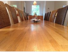 Ein langer Esstisch aus hellem Holz steht in einem hellen Raum mit geflochtenen Stühlen auf beiden Seiten. Im Hintergrund ist ein großes Fenster und eine Küchenzeile sichtbar, und auf dem Tisch steht eine flache Schale.