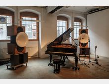 Ein heller Raum mit weiß verputzten Wänden und großen, bogenförmigen Fenstern. Im Vordergrund steht ein schwarzer Flügel, umgeben von Mikrofonen und zwei großen Lautsprechersystemen mit auffälligen weißen Hörnern.