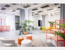 Heller, offener Studio- oder Coworking-Raum mit hohen Decken, Akustikpaneelen, großen Fenstern und grauen Vorhängen als Raumteiler. Im Vordergrund stehen Loungesessel und Hocker auf einem Teppich, daneben Pflanzen; im Hintergrund sind gelbe Tische und Stühle sowie rote Gitterregale und Stehtische zu sehen.