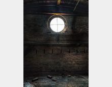 Ein verlassener Dachboden mit einer runden Fensteröffnung, die von verwittertem Mauerwerk umgeben ist. Der Raum ist dunkel, mit sichtbaren Dachbalken und Schutt auf dem Boden.