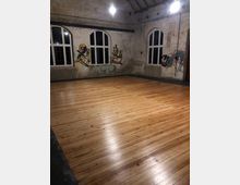 Ein leerer Raum mit einem Holzboden und sichtbarer Backsteinwand. Die Wände sind mit großen, bogenförmigen Fenstern und Graffiti verziert, während die Decke mit freiliegenden Holzbalken gestaltet ist.