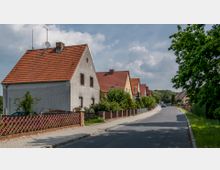 Straßenszene in einem Wohngebiet mit freistehenden Einfamilienhäusern, die rote Ziegeldächer und helle Fassaden haben. Links der Straße sind Gärten mit Hecken und Zäunen, rechts befindet sich ein bepflanzter Gehweg mit Bäumen.