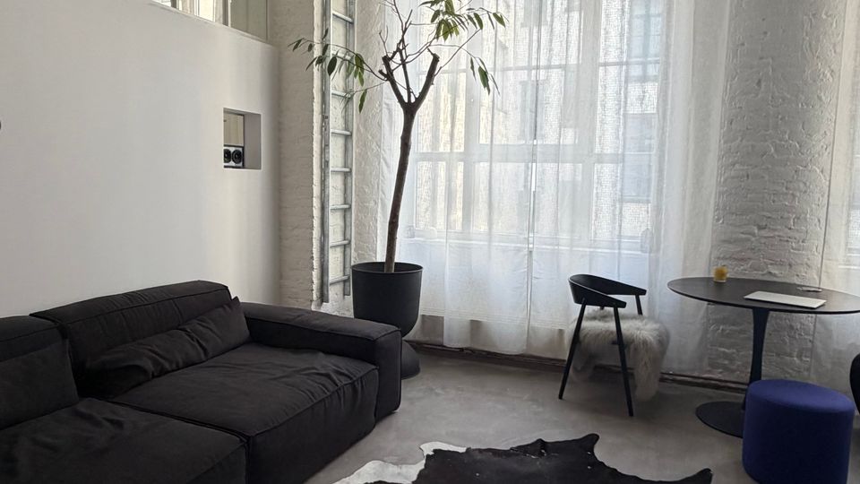 Heller, loftartiger Wohn- oder Arbeitsraum mit hohen Decken, weißen Backsteinwänden und großer Fensterfront mit transparenten Vorhängen. Im Vordergrund stehen ein schwarzes Ecksofa und ein schwarz-weißes Kuhfell auf Betonboden; daneben ein Topfbaum, ein kleiner Tisch mit Stuhl und ein blauer Hocker.
