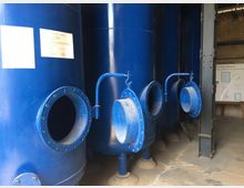 Große blaue Zylinder aus Metall mit offenen Inspektionsklappen in einer technischen Anlage, vermutlich Teil eines Wasseraufbereitungssystems. Im Hintergrund befindet sich eine Infotafel mit Text und Diagrammen.