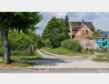 Ein schmaler, unbefestigter Weg führt entlang von Bäumen, Sträuchern und einem Drahtzaun zu einem Wohnhaus mit brauner Fassade und schrägem Dach. Rechts steht eine mit Graffiti verzierte Steinmauer neben einem Straßenschild mit der Aufschrift "Friedrich-Engels-Straße".