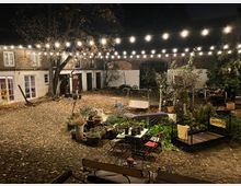 Ein gepflasterter Innenhof bei Nacht, beleuchtet durch Lichterketten. Umgeben von Backstein- und weiß getünchten Gebäuden mit großen Fenstern, befinden sich im Hof eine Mischung aus Pflanzen, Gartenmöbeln, Sitzgelegenheiten und Dekoration.