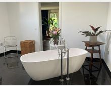 Modernes Badezimmer mit freistehender weißer Badewanne auf glänzendem schwarzen Fliesenboden und bodenstehender Chromarmatur. Neben der Wanne stehen Pflanzen und ein Blumenstrauß, dazu ein kleiner Holztisch; durch eine offene Tür ist ein angrenzender Raum mit Fensterblick ins Grüne zu sehen.