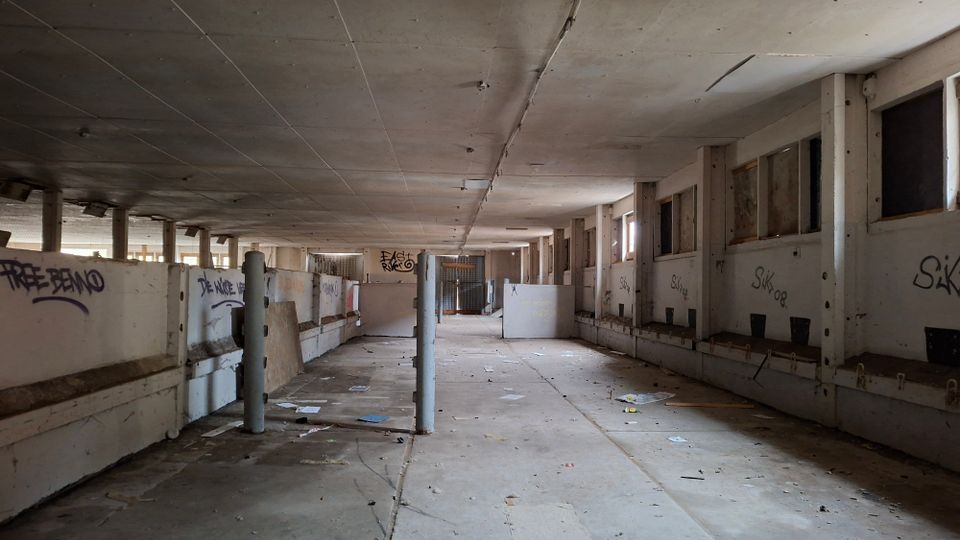 Innenansicht einer großen, verlassenen Industriehalle mit niedriger Betondecke, Stützpfeilern und seitlichen Trennwänden mit Graffiti. Der lange Gang führt zu einem vergitterten Bereich am Ende; auf dem Boden liegen verstreute Abfälle und einzelne Platten.