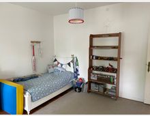 Zimmer eines Kindes mit einem weißen Einzelbett, das mit einer blauen Bettdecke mit Sternenmuster bezogen ist. Rechts steht ein Holzregal mit Spielzeug, Büchern und einer kleinen Stereoanlage. Wanddekorationen und helle Wände vervollständigen den Raum.