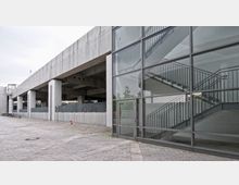 Ansicht eines modernen Gebäudekomplexes mit Sichtbeton-Elementen, einer überdachten Passage sowie einer verglasten Treppenhausfassade rechts im Bild. Der gepflasterte Vorplatz ist weitläufig und leer, mit Fahrrädern im Hintergrund.