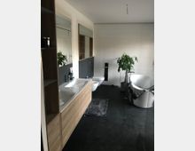 Modernes Badezimmer mit großformatigen hellen Wandfliesen und dunklen Bodenfliesen, Holz-Waschtisch mit Doppelwaschbecken und großem Spiegelschrank. Rechts stehen eine freistehende Badewanne mit Handtuch, ein bodenstehender Wasserhahn und eine große Topfpflanze.