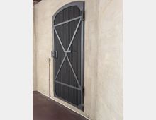 Schwarze Metalltür mit einem gewölbten oberen Rand und diagonalen Verstrebungen, eingebaut in eine beige Betonwand; der Boden besteht aus dunkelbraunem Material.