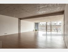 Großer, leerer Raum mit Betonwänden und -decke, hellem Holzfußboden und bodentiefen Fenstern, die einen Blick auf eine Außenterrasse und Bäume bieten. Die minimalistische Einrichtung umfasst einzelne Pflanzen und dezente Lichtquellen.