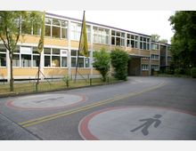 Zweistöckiges Schulgebäude aus gelbem Backstein mit großen rechteckigen Fenstern, flankiert von Bäumen und umgeben von einem niedrigen Zaun. Im Vordergrund befindet sich eine Asphaltfläche mit eingezeichneten Fußgängermarkierungen und gelben Fahnen.