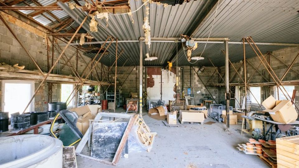 Ein altes, heruntergekommenes Lagerhaus mit Wellblechdach, freiliegenden tragenden Metallstreben und unverputzten Betonwänden. Der Raum ist gefüllt mit verschiedenen Materialien, Kartons, Werkzeugen und verstreuten Gegenständen unter einem teilweise beschädigten Dach.