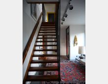 Moderne Wohnraumgestaltung mit einer Holztreppe mit offenen Stufen, die nach oben zu einem Geländer aus schmalen schwarzen Streben führt. Der Boden ist mit einem roten, gemusterten Teppich bedeckt, daneben stehen ein Sofa und eine Designerlampe; die weißen Wände sind mit Kunstwerken und Dekoration akzentuiert.