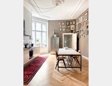 Bright open-plan kitchen and dining area with high ceilings, ornate ceiling molding, and large multi-pane windows. A long dining table sits on herringbone wood floors beside modern kitchen cabinets, a red runner rug, a stainless-steel fridge, and a gallery wall of framed artwork.