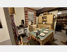 Ein retrostiliger Küchenraum mit einem Ofen, einem Kühlschrank, einer Anrichte und einem gedeckten Tisch mit Tellern. Im Hintergrund sind Vorhänge mit einem gemusterten Stoff und ein Holzregal zu sehen.