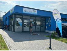 Ein einstöckiges, modernes Gebäude mit blauer Fassade und großen Glasfenstern, gekennzeichnet als "Rezeption" des "Stream Camp". Vor dem Eingang stehen Fahrräder, eine Person betritt das Gebäude, und rechts ist ein Werbebanner des Camps zu sehen. Im Vordergrund führt ein gepflasterter Weg zum Eingang.