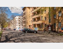 Straßenszene in einem Wohngebiet mit mehrstöckigen Wohnhäusern aus gelbem Putz und Backsteindetails. Die Häuser sind von jungen Bäumen gesäumt, es sind geparkte Autos, Fahrräder und mit Graffiti versehene Wände zu sehen, bei sonnigem Wetter.