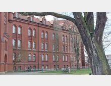 Backstein-Gebäude im neogotischen Stil mit mehreren Stockwerken und charakteristischen Rundbogenfenstern, im Vordergrund Bäume, eine Bank und ein Fahrradständer in einer parkartigen Umgebung.