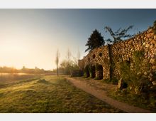 Eine Landschaft mit einer gepflasterten Steinmauer auf der rechten Seite, die von Pflanzen teilweise überwachsen ist, und einem Weg, der daran vorbeiführt. Im Hintergrund stehen Bäume und Gebäude, während die Szene in warmes, goldenes Licht der untergehenden Sonne getaucht ist.