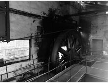 Schwarz-Weiß-Aufnahme eines industriellen Innenraums mit einer großen, historischen Schwungradmaschine an einer abgenutzten Wand. Metallstege und Geländer durchziehen den Raum, während Oberlichter gedämpftes Licht einfallen lassen.