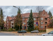 Straßenansicht eines mehrstöckigen Wohnhauses aus rotem Ziegelstein mit Satteldach und verzierten Balkonen, umgeben von herbstlichen Bäumen. Im Vordergrund stehen geparkte Autos, Straßenschilder und eine Laterne.