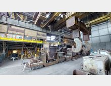 Industriehalle mit großen Kranhaken im Vordergrund, Schmelzöfen auf Schienenwagen und Metallkonstruktionen im Hintergrund. Die Wände bestehen aus Backstein und Metall, die Halle ist durch gelbe Geländer unterteilt, und Deckenkrananlagen sind sichtbar.