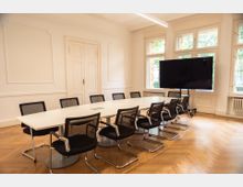 Ein moderner Besprechungsraum mit einem weißen Konferenztisch, umgeben von schwarzen Bürostühlen. Der Raum hat helle Wände mit dezenten Wandverzierungen, große Fenster, durch die Tageslicht einfällt, und einen großen Bildschirm auf einem Rollständer.