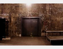 Industrieähnlicher Raum mit abgenutzter, graffitibeschmierter Wand. Zentral befindet sich eine metallene Tür mit der Aufschrift „Technik-Lager“, daneben stehen ein Metallgestell und eine aufgerollte rote Kabeltrommel.