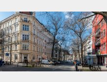 Straßenszene in einer Stadt mit Altbauten. Auf der linken Seite steht ein beigefarbenes Gebäude mit dekorativen Fassadenelementen, rechts ein rotes Mehrfamilienhaus, Bäume ohne Laub säumen die Straße, auf der Autos parken und vereinzelt Menschen unterwegs sind.