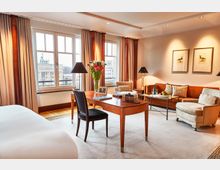 Helles, elegantes Hotelzimmer mit großem Schreibtisch aus Holz, gemütlicher Sitzecke und einem großen Fenster mit Blick auf das Brandenburger Tor. Die Einrichtung umfasst beige Vorhänge, warme Holzakzente, zwei gerahmte Bilder an der Wand und dezente Beleuchtung.