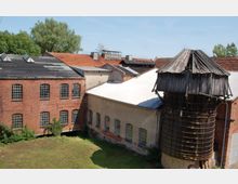 Ehemaliger Industriekomplex mit Backstein- und verputzten Gebäuden, großen Sprossenfenstern und einem baufälligen Holzsilo; im Vordergrund ein grasbewachsener Innenhof, im Hintergrund teils zerstörte Ziegeldächer und Bäume.