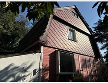 Ein zweistöckiges Haus mit steilem Satteldach, verkleidet mit rautenförmigen Dachschindeln und roter Holzfassade. Es liegt inmitten von Bäumen, mit sichtbaren Fenstern und einem umliegenden Gartenbereich.