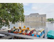 Ein Flussufer mit mehreren bunt bemalten Hausbooten, die an einem hölzernen Steg festgemacht sind. Im Hintergrund ein großes, historisch wirkendes Backsteingebäude mit einem modernen Dachausbau sowie Bäume und weitere Wasserfahrzeuge.