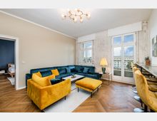 Helles Wohnzimmer mit Fischgrätparkett, großem Ecksofa in Dunkelblau sowie gelbem Sessel und Hocker um einen niedrigen Couchtisch auf Teppich. Hohe Fenster und eine Balkontür mit Blick nach draußen, dazu eine Barzeile mit gepolsterten Hockern an der rechten Wand und eine Deckenleuchte.