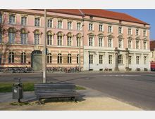 Historisches Gebäude mit rosa und cremefarbener Fassade, verzierten Fenstern und einem roten Ziegeldach, umgeben von geparkten Fahrrädern. Im Vordergrund stehen eine Holzbank und ein Mülleimer an einer Kreuzung.