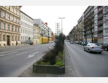 Straßenszene in einer urbanen Umgebung mit mehrspuriger Fahrbahn, flankiert von historischen und modernen Wohn- und Geschäftsgebäuden. In der Mitte der Straße befindet sich ein mit Pflanzen und Metallgittern gestalteter begründer Bereich, während Autos am Straßenrand und in Bewegung zu sehen sind.